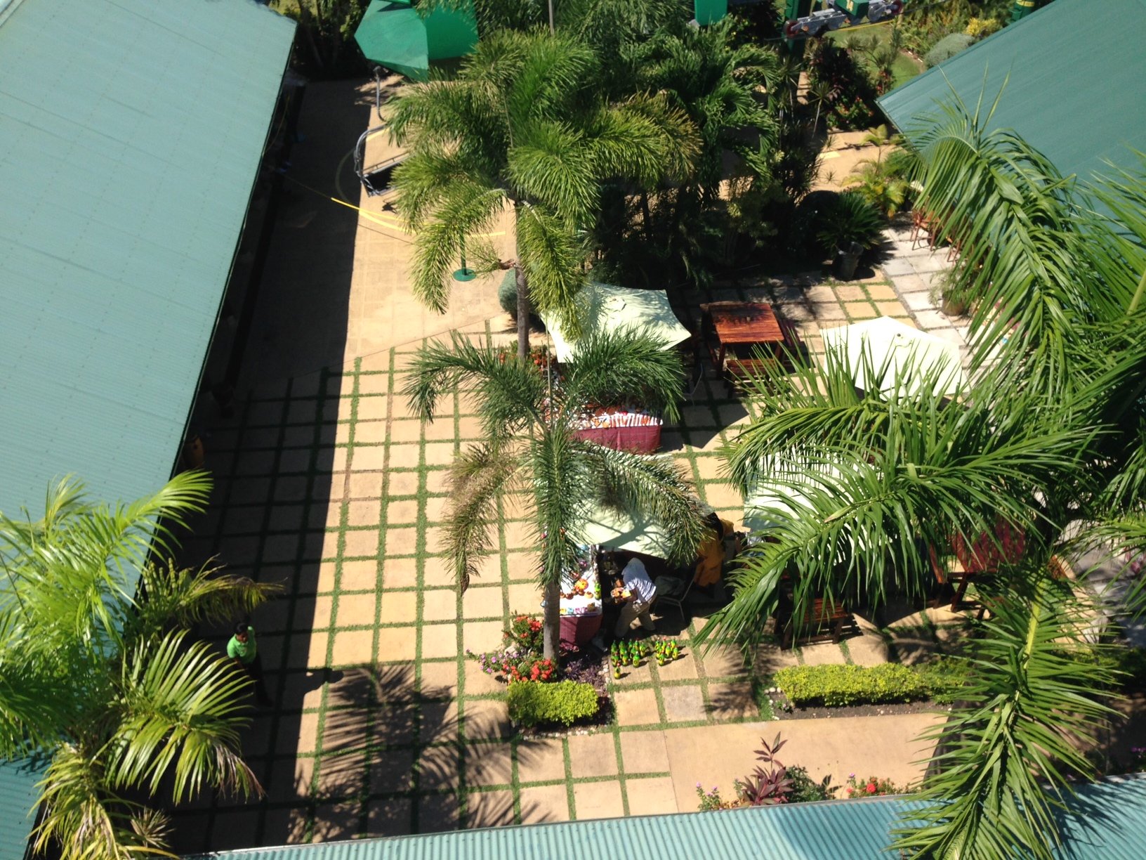 Overhead view of a paved courtyard area with a grid pattern, featuring small square garden beds, palm trees, and outdoor seating with white umbrellas.