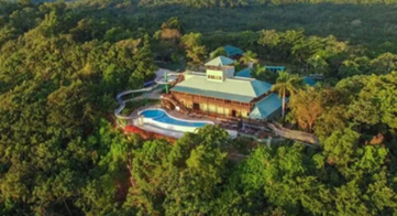 Aerial view of a large multi-story building with a green roof, a swimming pool, and surrounding decks, nestled within dense tropical rainforest.