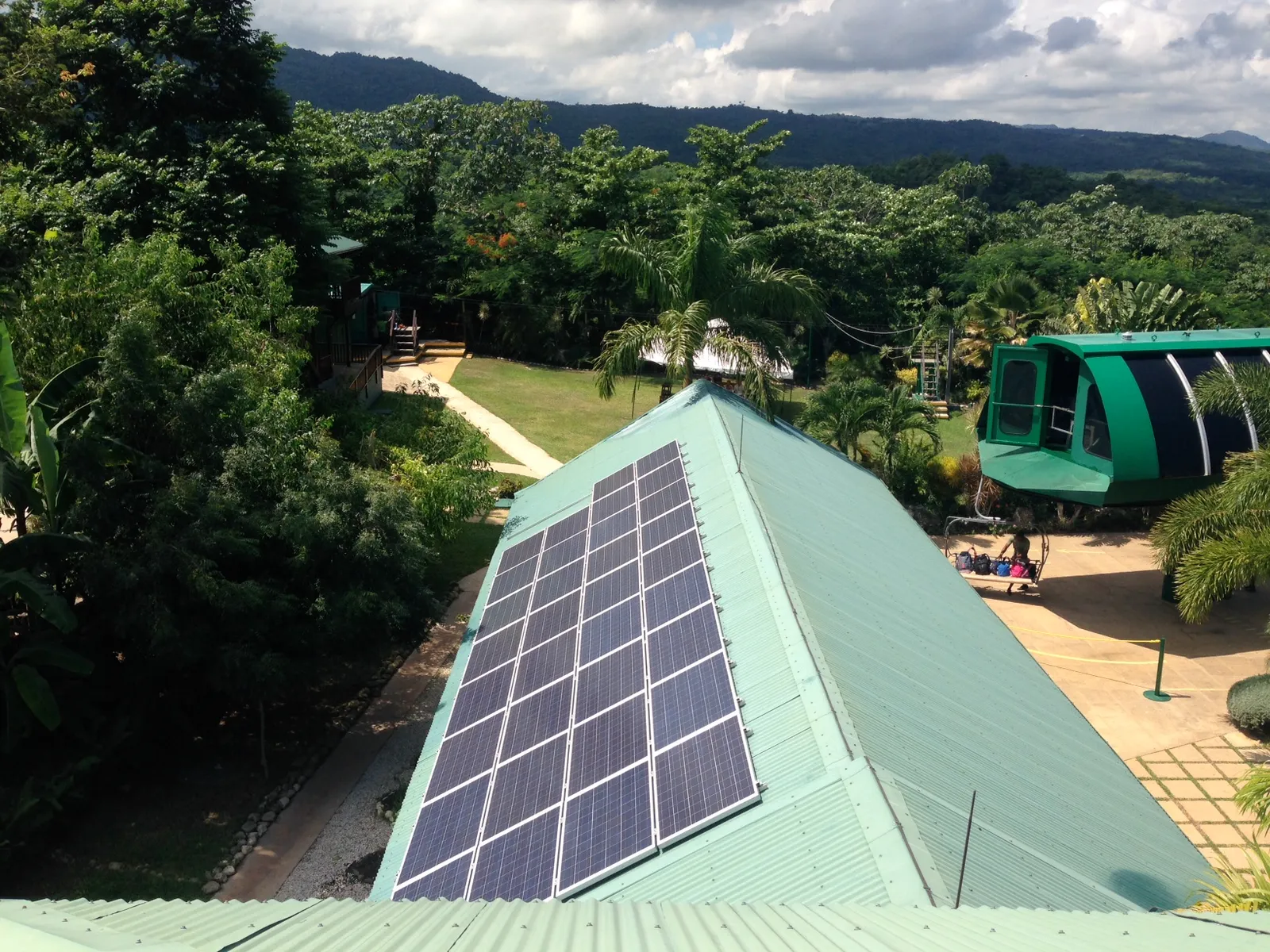 High-angle view of Mystic Mountain’s green-roof lodge and pool perched in dense rainforest, with lift station and sea beyond.