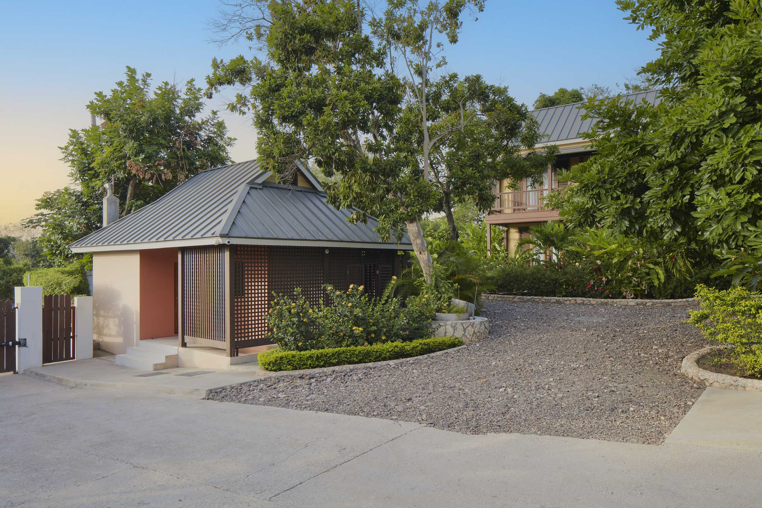 Exterior view of the entrance area with a small structure and gate, leading to a gravel driveway. Two cottages are visible through the trees in the background.
