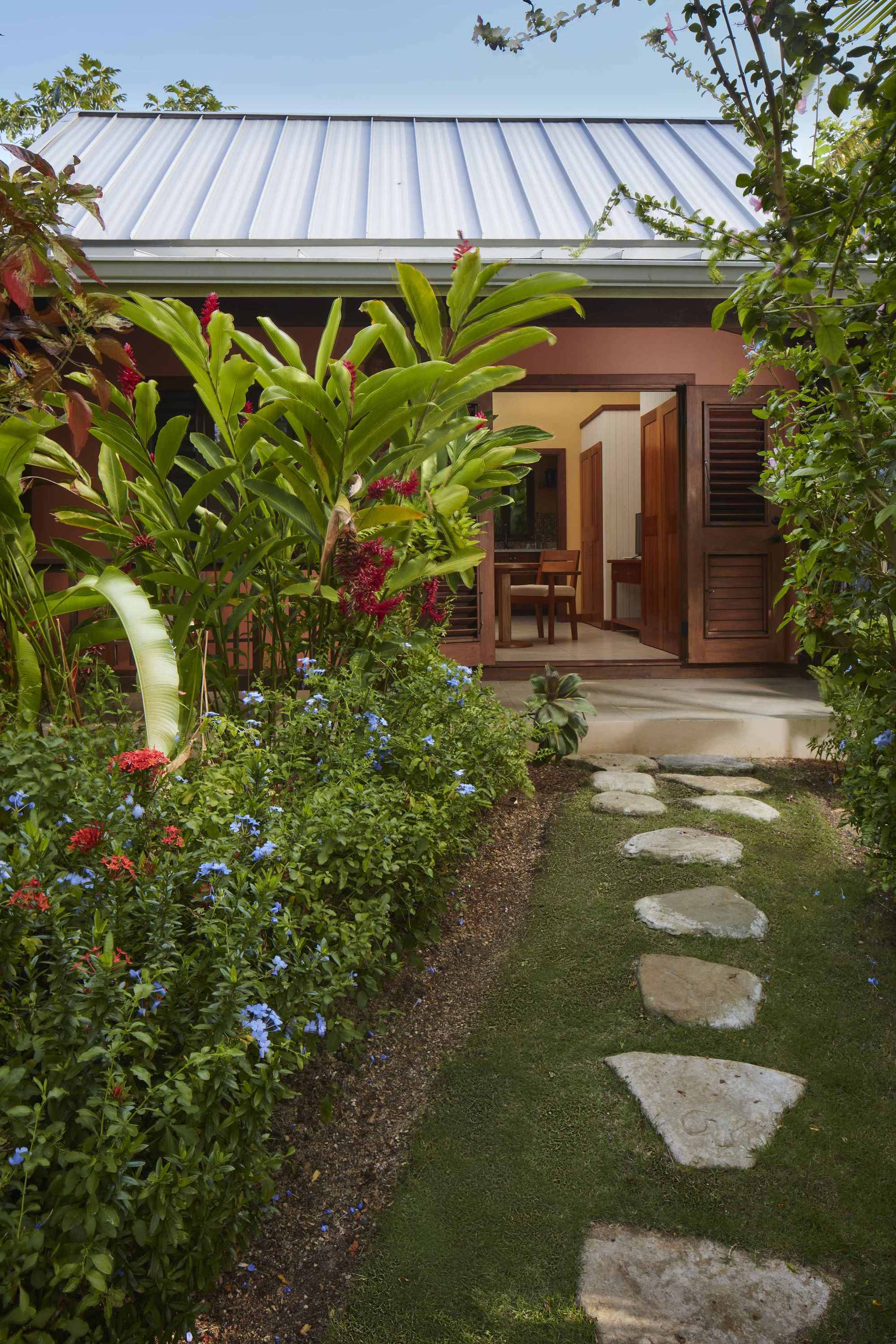 A stone path leads through a dense garden of flowering blue and red tropical plants toward the open doorway of a cottage.