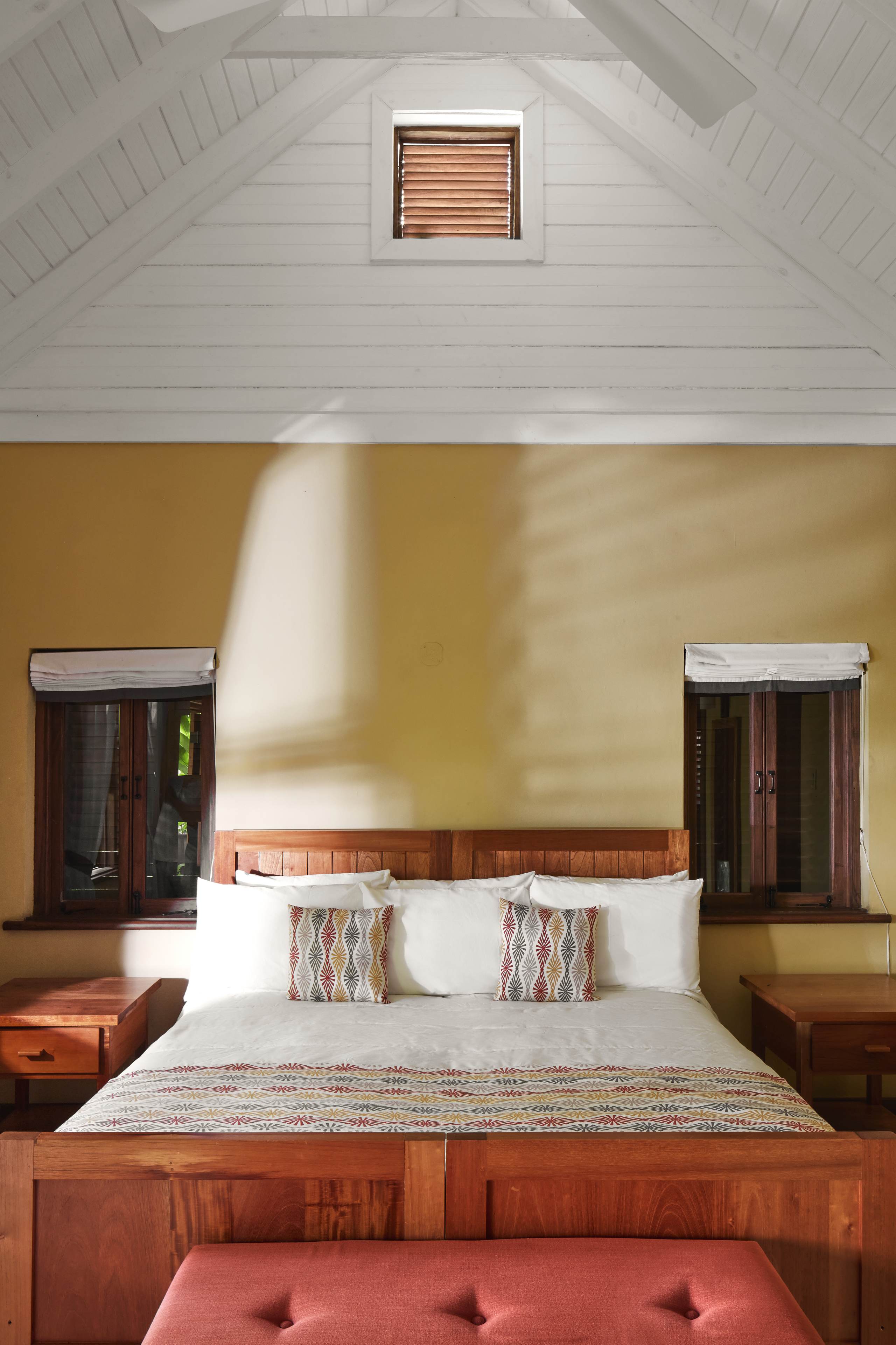 Interior view of a cottage bedroom featuring a wooden bed frame, white bedding, and colorful accent pillows against a warm yellow wall. The ceiling is white and vaulted with a small window at the peak.