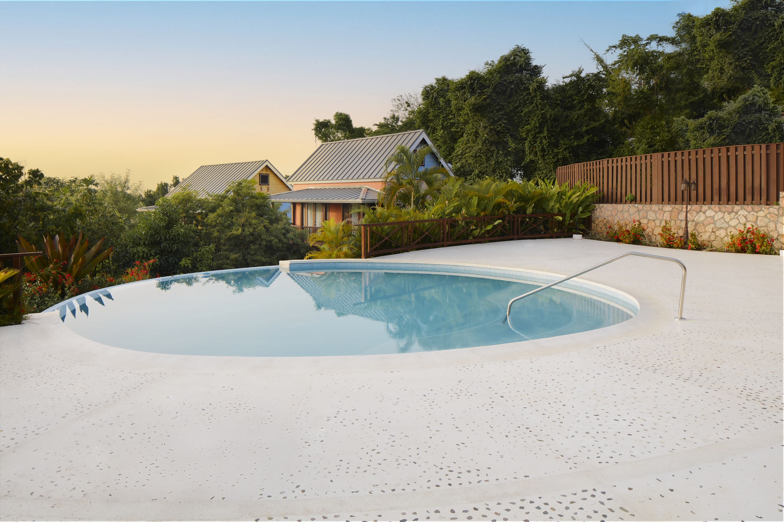 Wide shot of a kidney-shaped, white-rimmed swimming pool with a metal handrail, surrounded by a light-colored patio deck, with two cottages visible among green trees in the background.