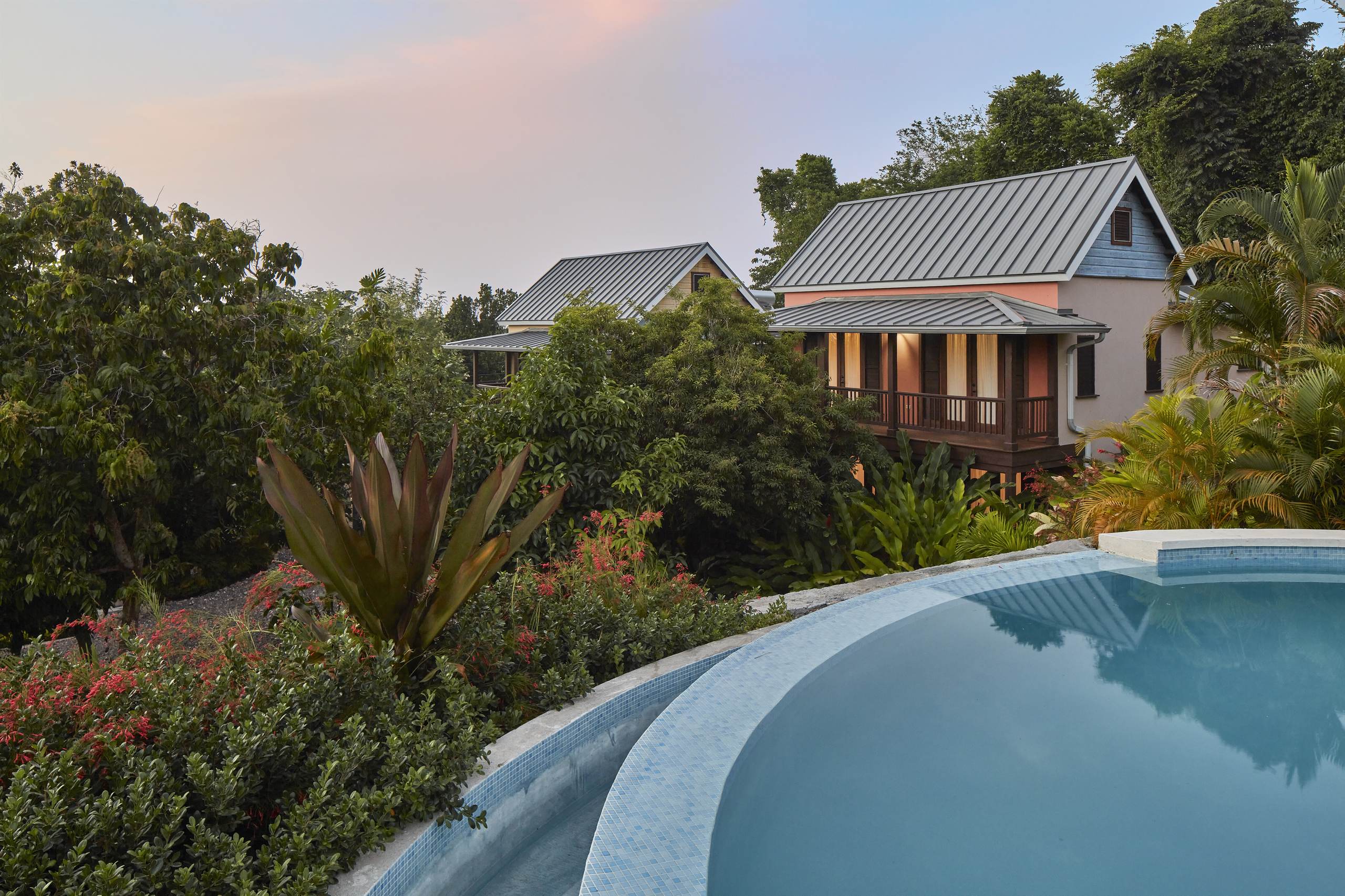 View of two hillside cottages with dark metal roofs, nestled in lush tropical vegetation, overlooking the curved edge of an infinity pool at sunset.
