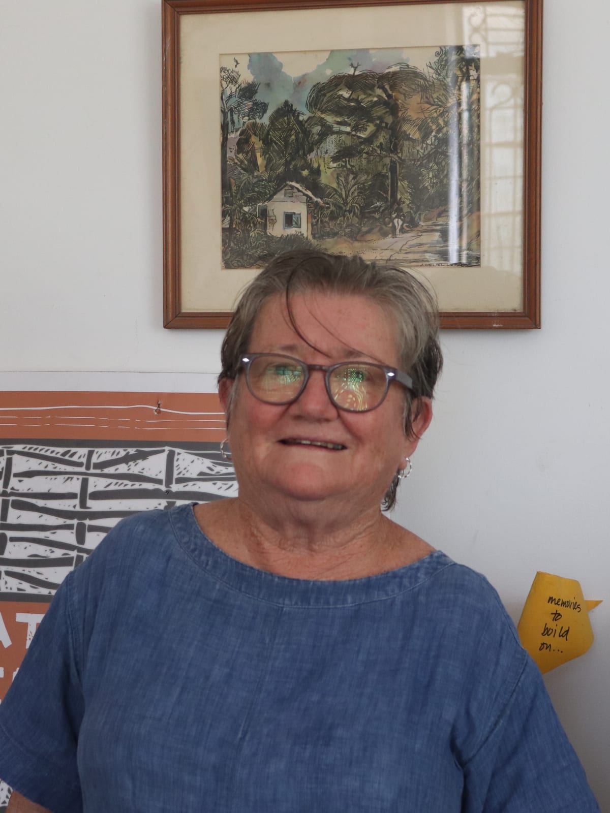 portrait indoors, smiling in denim-blue dress, framed artwork and studio posters on the wall.