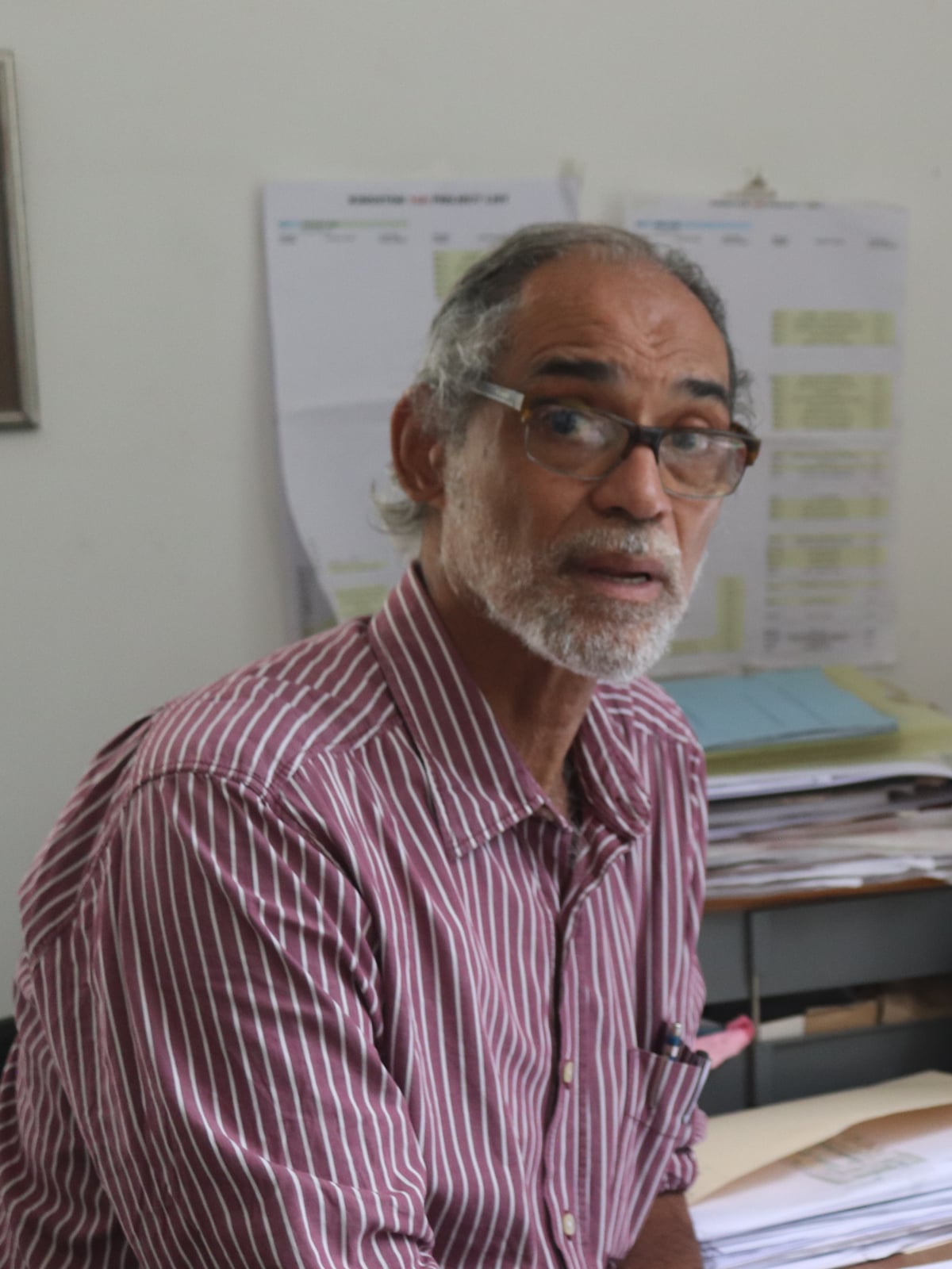 portrait at desk in striped burgundy shirt and glasses, folders and pinned schedules behind.
