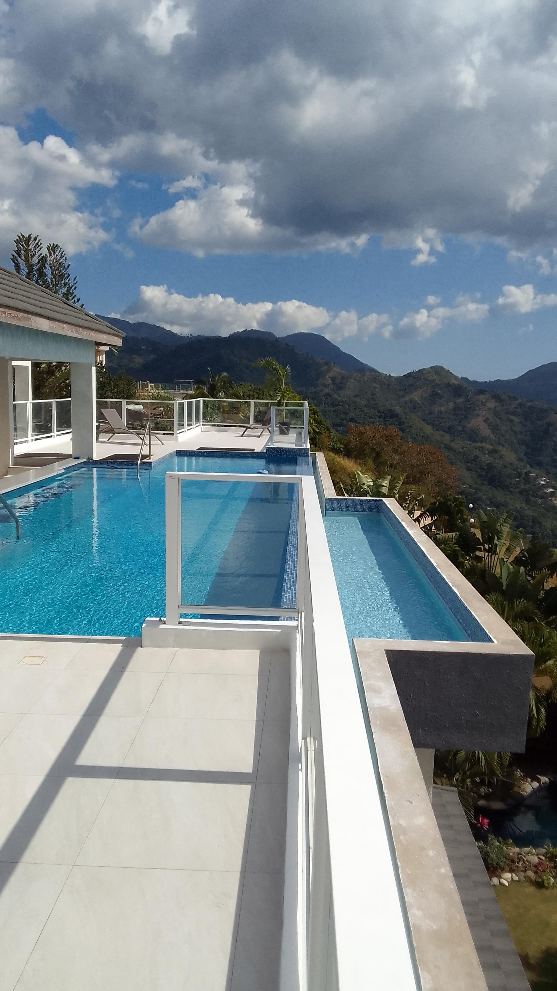Infinity pool with glass railing on a patio overlooking a vast, lush mountain range and valley under a bright blue sky with white clouds. A smaller secondary pool is adjacent to the main pool.