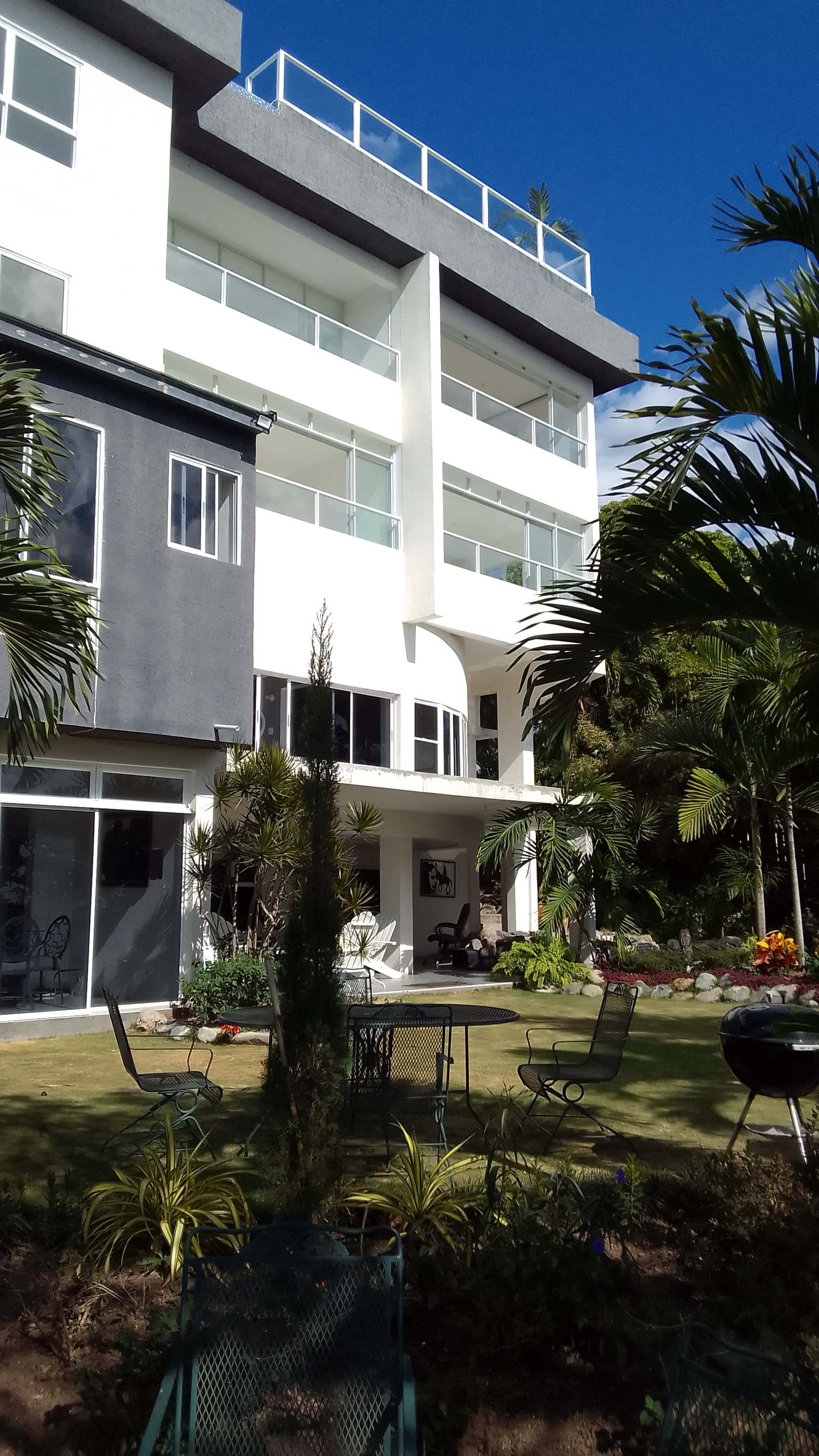Low-angle view of the modern white and gray apartment building's facade, showing multiple levels of glass-railed balconies and a ground-floor patio area. The foreground features a manicured lawn with garden furniture and tropical plants.
