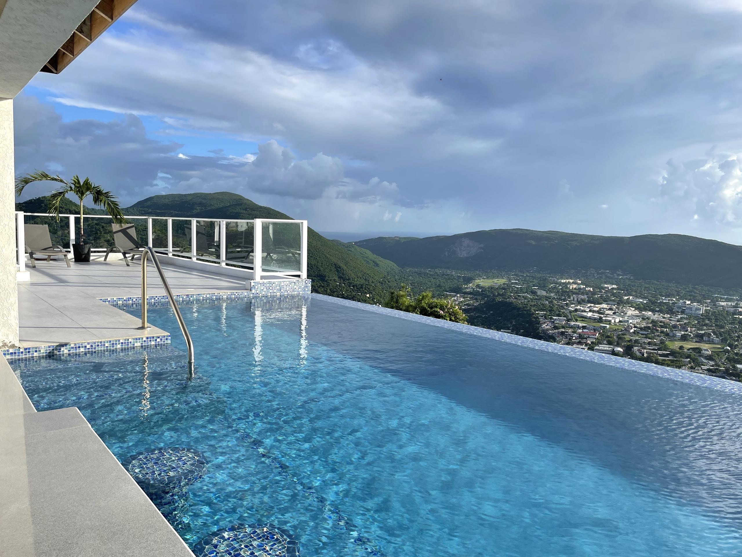 Edge of an infinity swimming pool looking out over a wide, hilly landscape with a town or city nestled in the valley below. The sky is partly cloudy with patches of blue.
