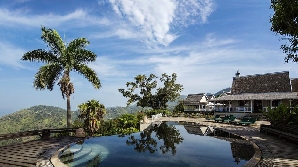curved infinity-edge pool reflecting palm and sky with mountain backdrop and verandah buildings beyond.