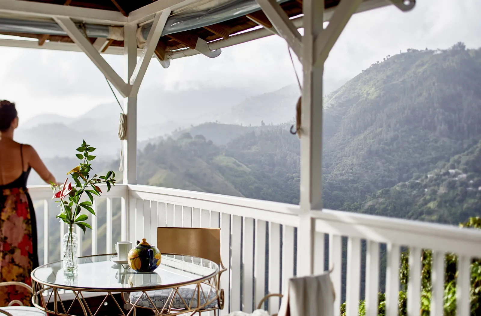 verandah breakfast table with teapot, cup and flowers overlooking cloud-washed Blue Mountains ridges, person leaning on rail.