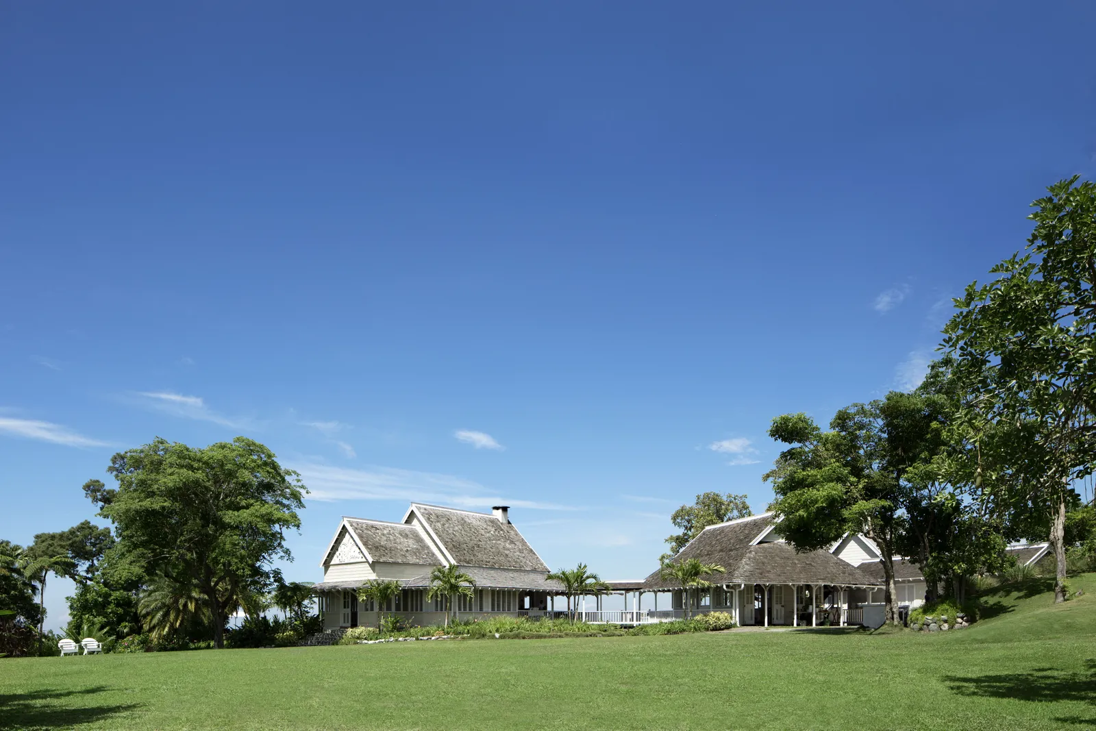pair of timber great houses with louvered shutters framed by palms on a bright open lawn under wide blue sky.