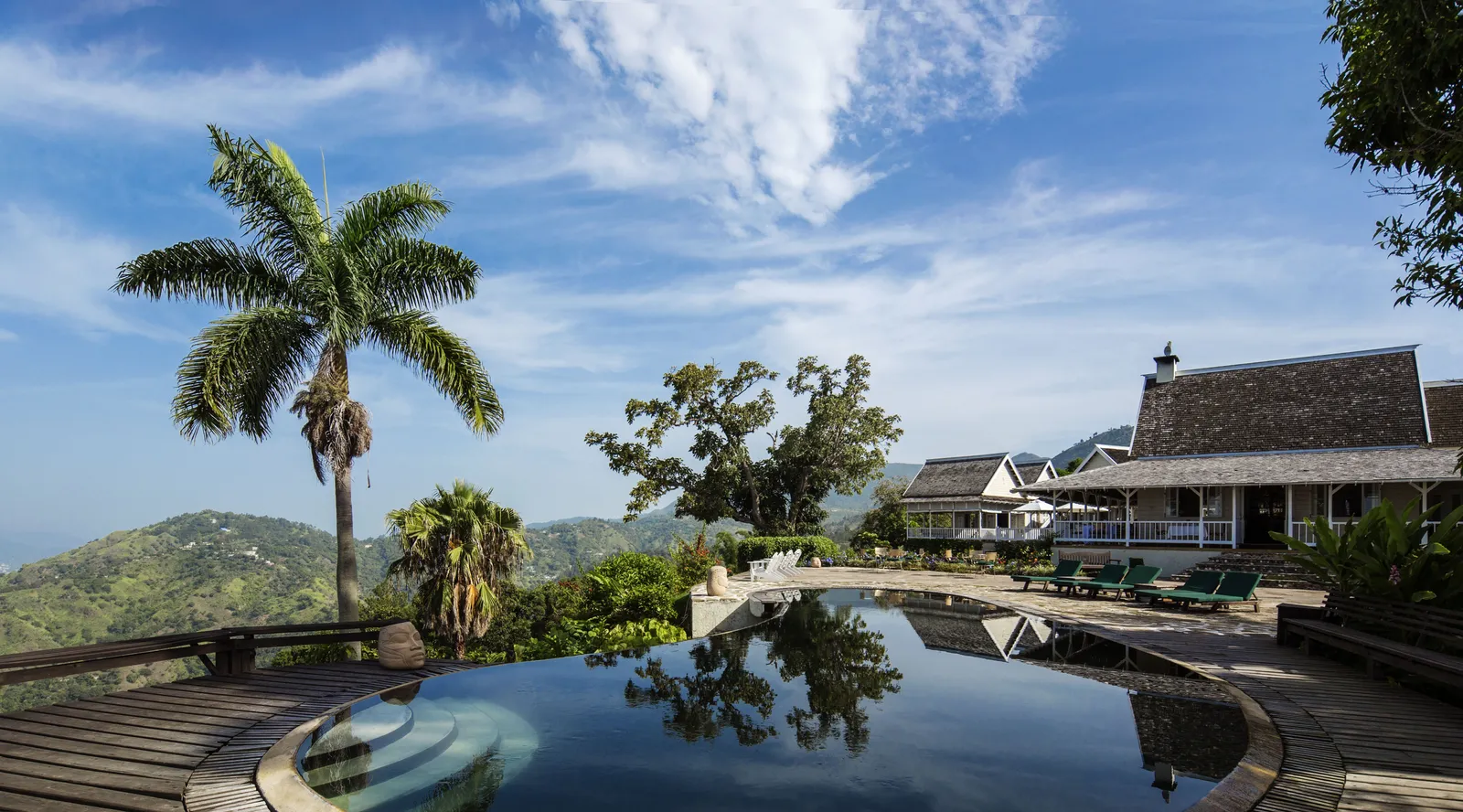curved infinity-edge pool reflecting palm and sky with mountain backdrop and verandah buildings beyond.