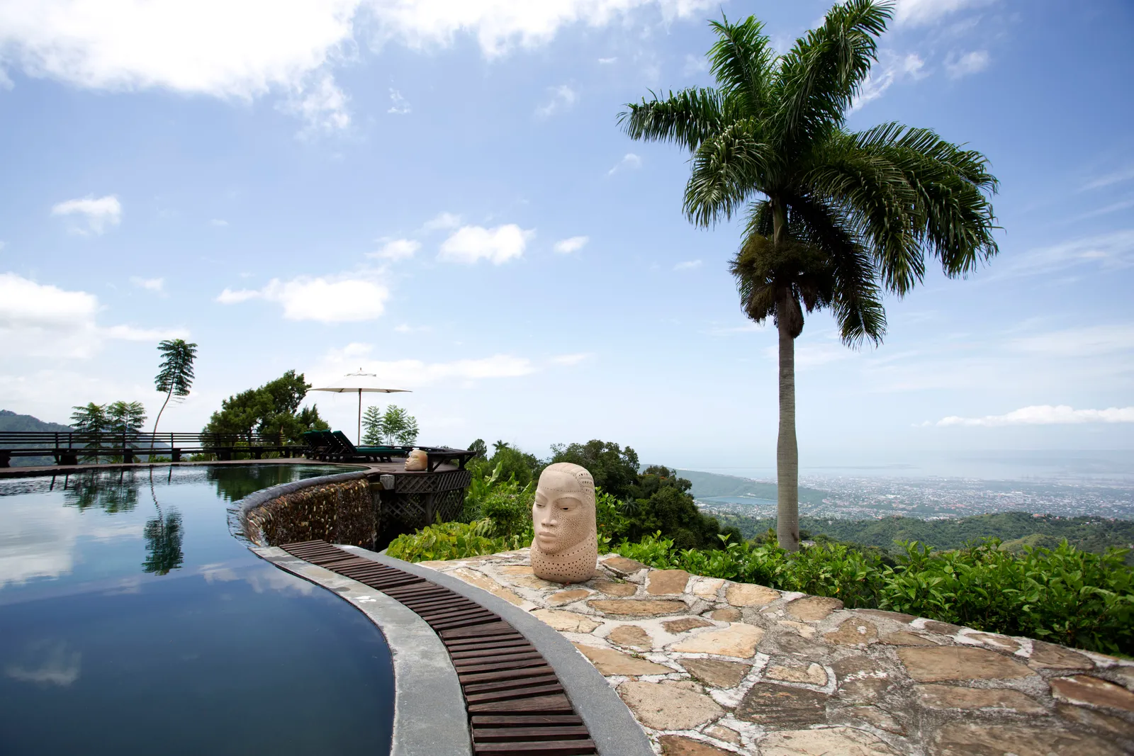 pool terrace with stone paving, carved head sculpture and tall palm overlooking Kingston and the coast far below.