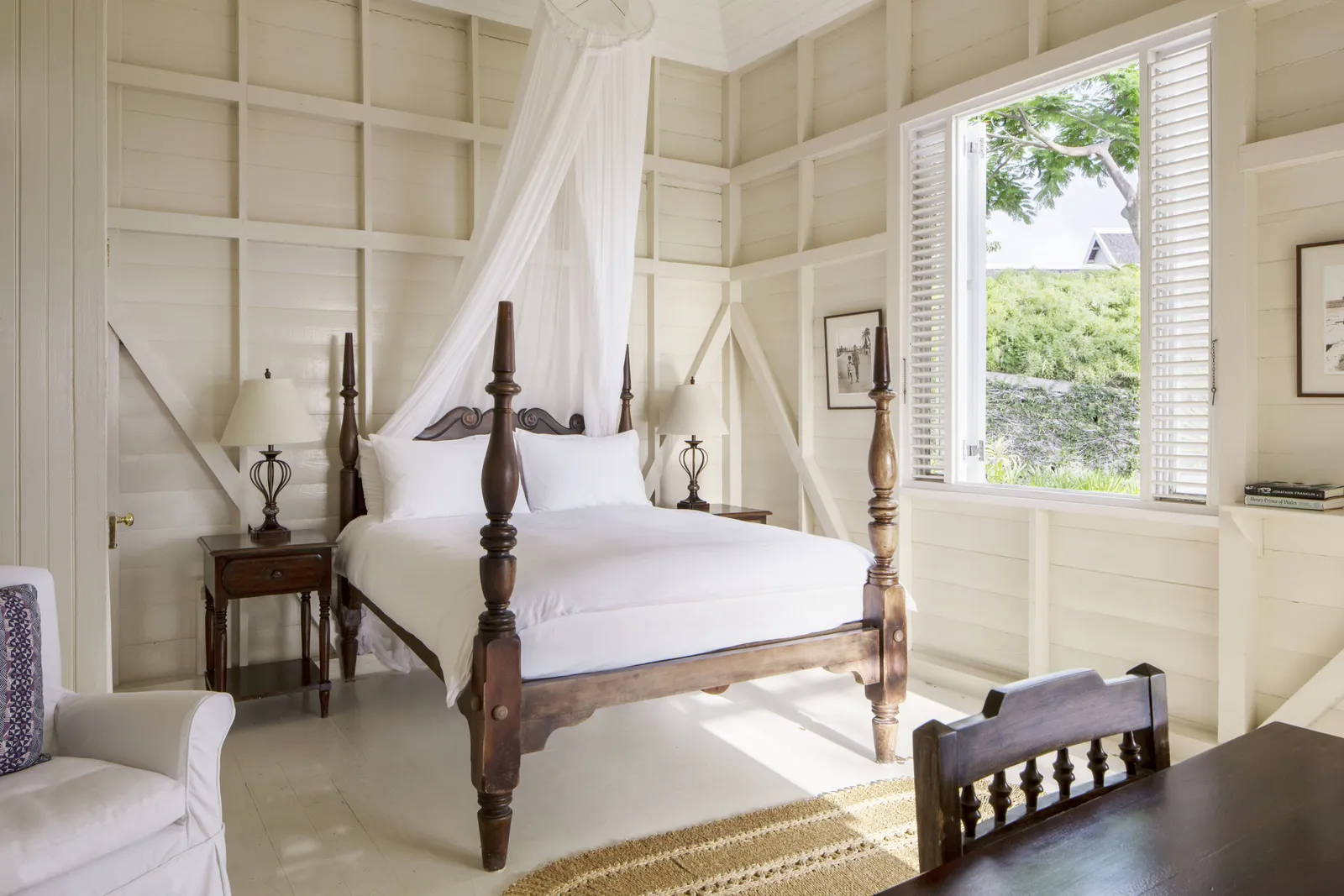 airy cottage bedroom with four-poster bed draped in mosquito netting, white timber walls and open louvered window to garden.