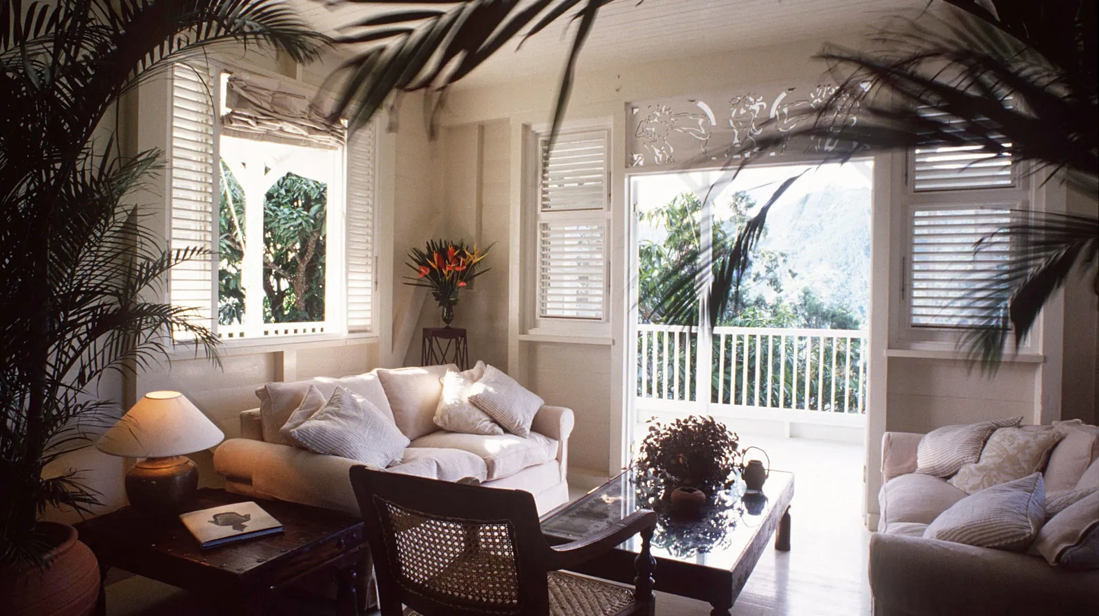 sitting room framed by palms, soft sofas and glass coffee table, louvered windows and open door to balcony facing the hills.