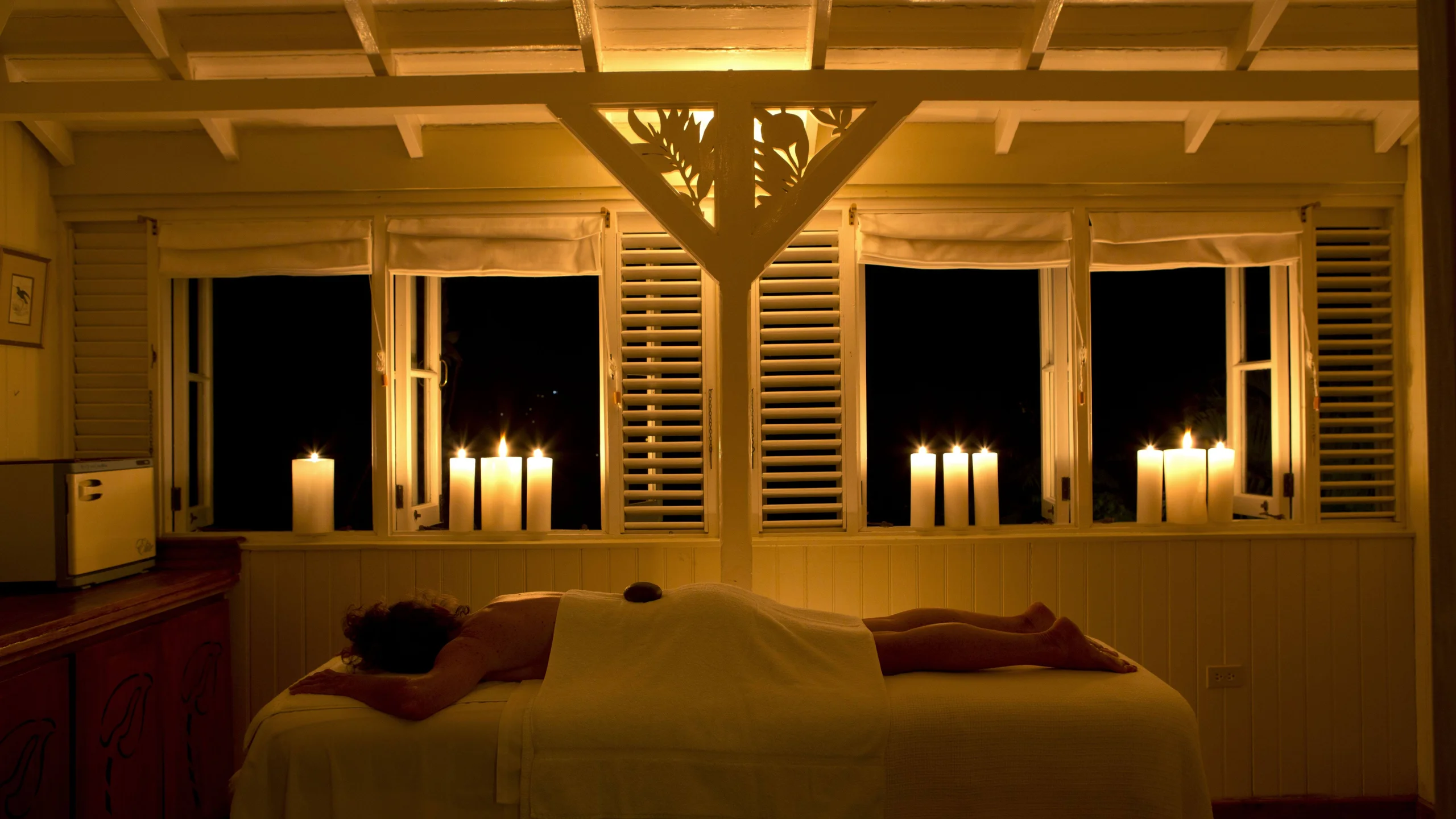 candlelit spa room at night, guest lying face down for treatment beneath louvered windows with rows of tall candles.